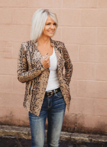 A woman with platinum blonde hair wears a snake-print blazer and jeans, standing with a relaxed demeanor against a textured pink wall.
